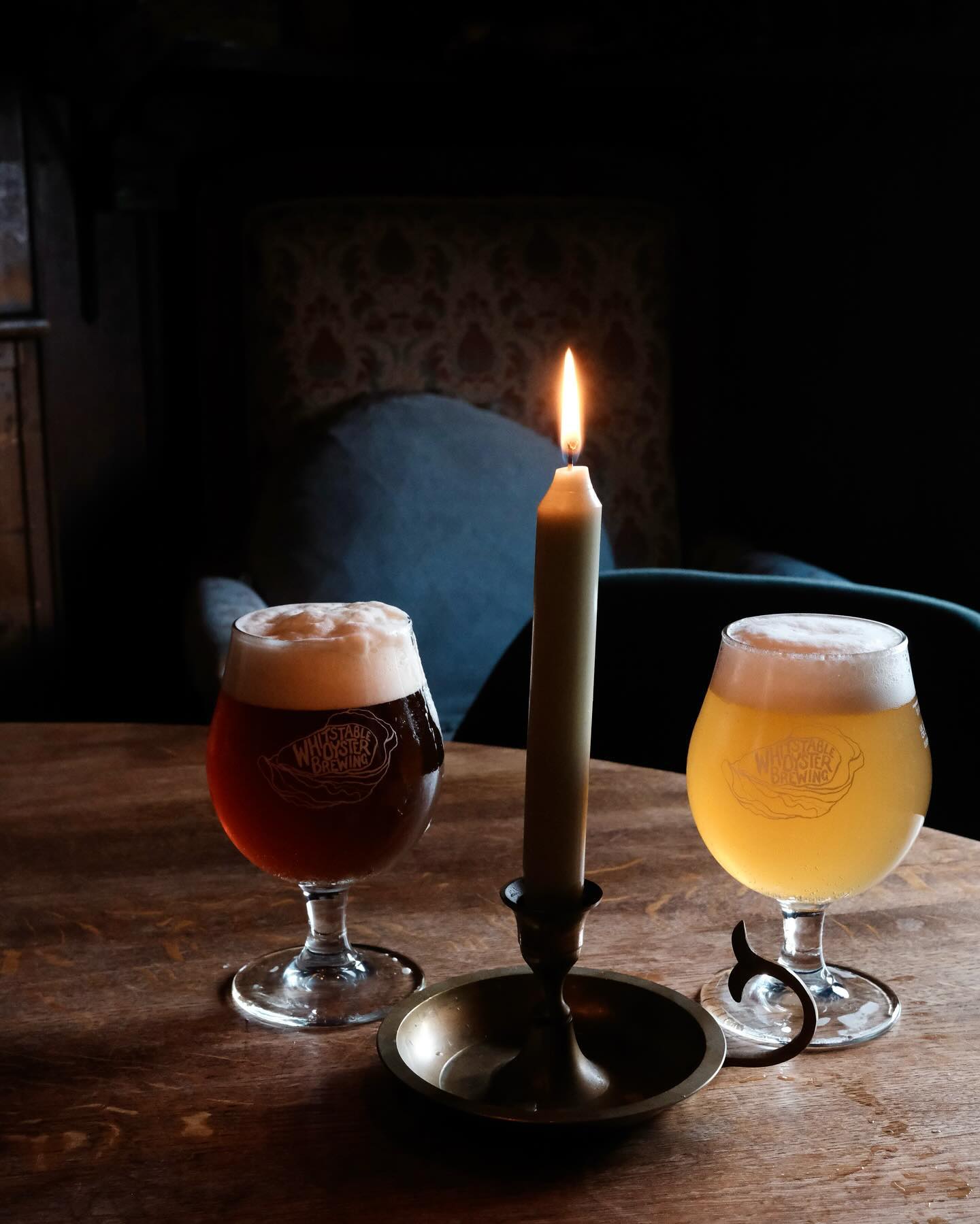 A glass of Whitstable Oyster Brewing 'Teak' 5.2% Green Hop ESB featuring fresh Goldings hops & a glass of 'Brass' 5.2% Green Hop Pale featuring fresh Ernest hops sit on a wooden table, with a lighted candle between them.